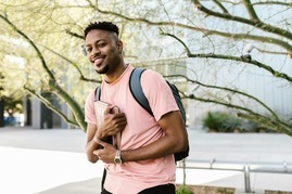 Smiling student outdoors holding a book labeled 'Law,' wearing a pink t-shirt and a backpack, standing in a natural setting with trees and modern architecture in the background.